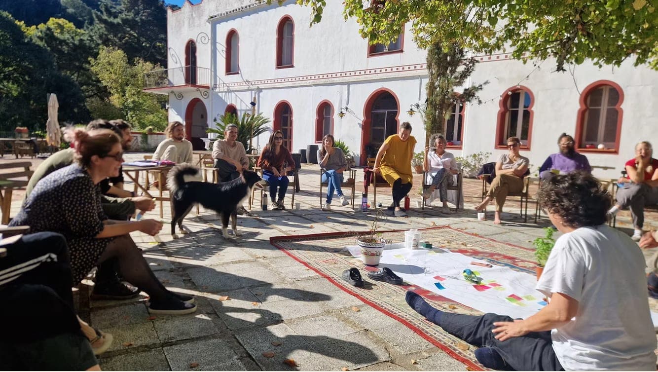 A group sitting in a circle in the courtyard of a white house, with a dog and large sheets of paper on the ground